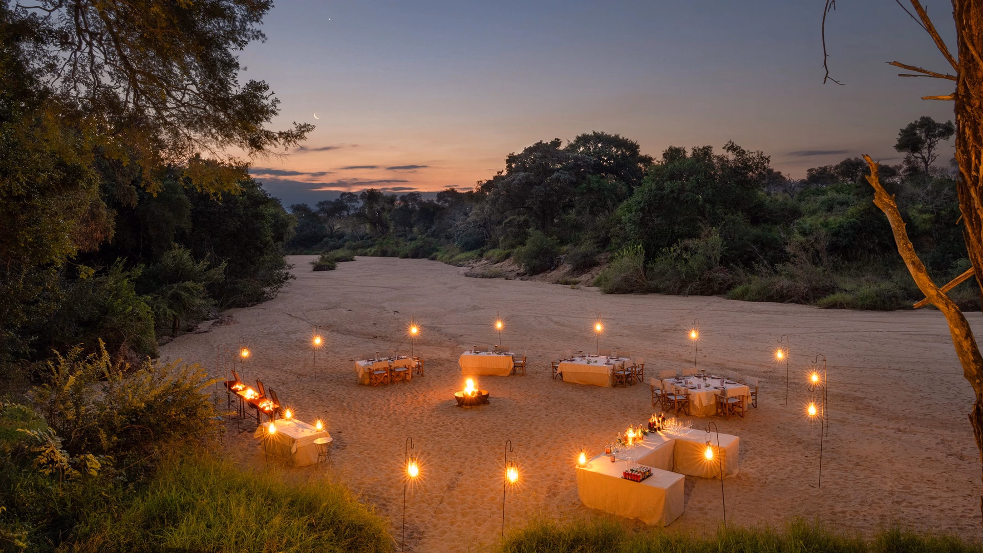 A lantern-lit bush dinner on a dry riverbed at Seseka Tented Camp, with tables set around a fire under a crescent moon at dusk.