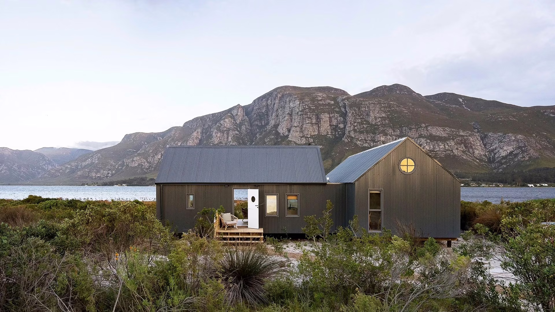 Coot Club Boathouse, a modern charcoal-clad cottage with corrugated iron roofs set among coastal fynbos beside a lagoon, with rugged sandstone mountains rising behind.